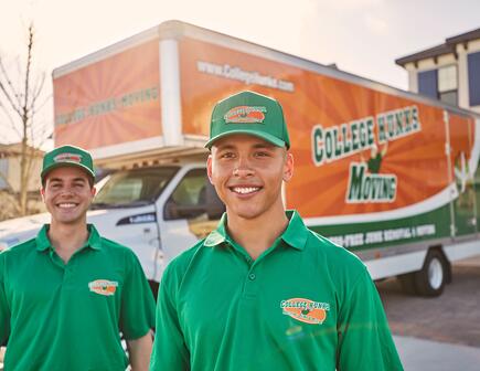 Two men wearing green College Hunks shirts, standing in front of a moving truck.