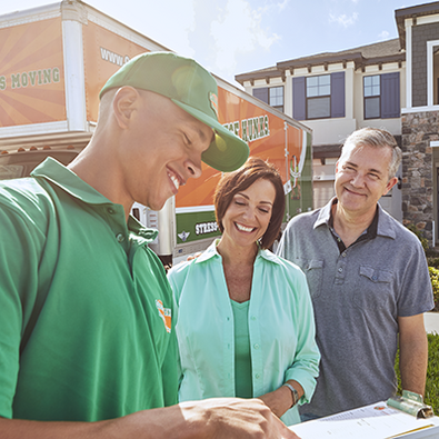 A mover standing with two clients in front of an orange and green moving truck titled "College Hunks Hauling Junk & Moving" 